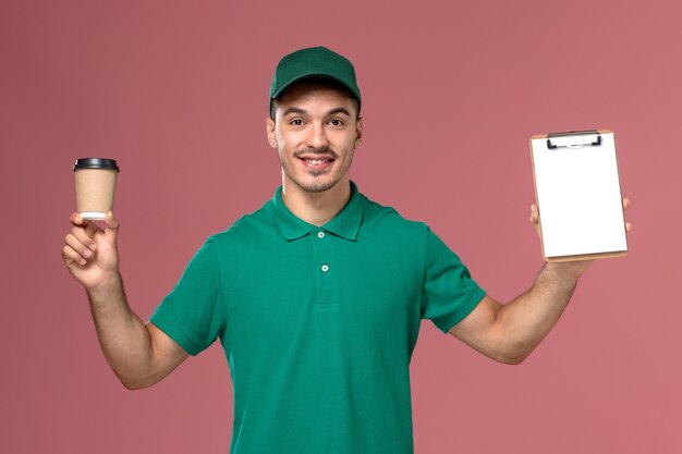 Mensajero masculino de vista frontal en uniforme verde sosteniendo la taza de café y el bloc de notas sobre fondo rosa