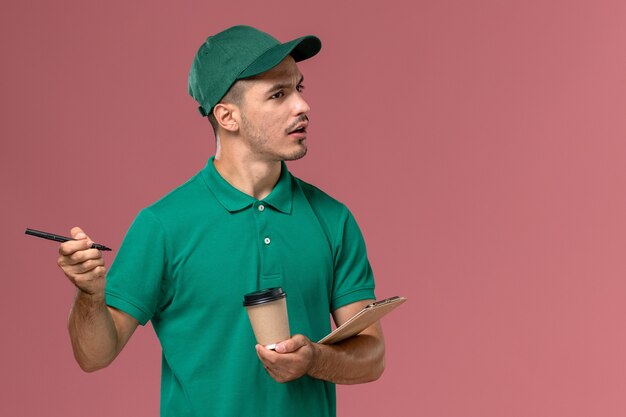 Mensajero masculino de vista frontal en uniforme verde sosteniendo la taza de café de entrega y el bloc de notas escribiendo notas en el escritorio rosa claro