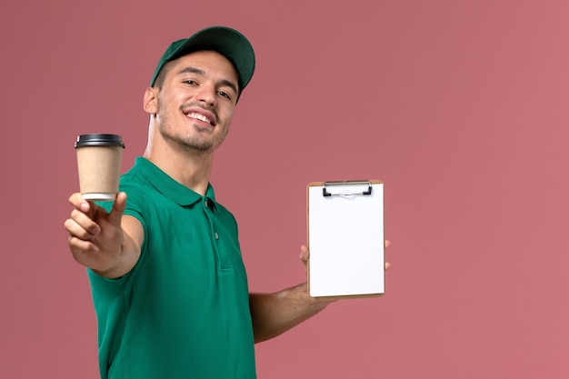 Mensajero masculino de vista frontal en uniforme verde sosteniendo la taza de café de entrega y el bloc de notas sonriendo en el escritorio de color rosa claro