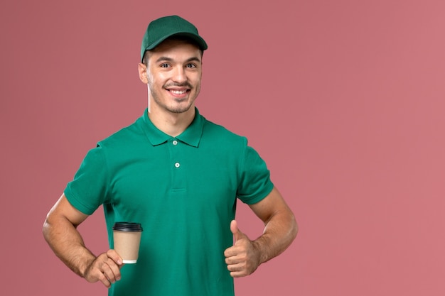 Foto gratuita mensajero masculino de vista frontal en uniforme verde sosteniendo la taza de café de entrega con una leve sonrisa en el trabajador de fondo rosa