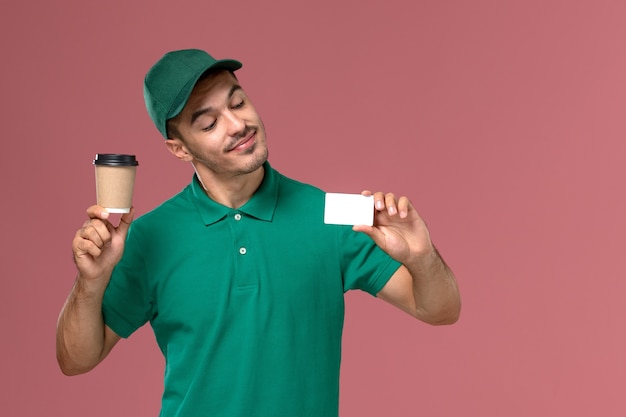 Mensajero masculino de vista frontal en uniforme verde sosteniendo la taza de café de entrega con tarjeta blanca en el escritorio rosa