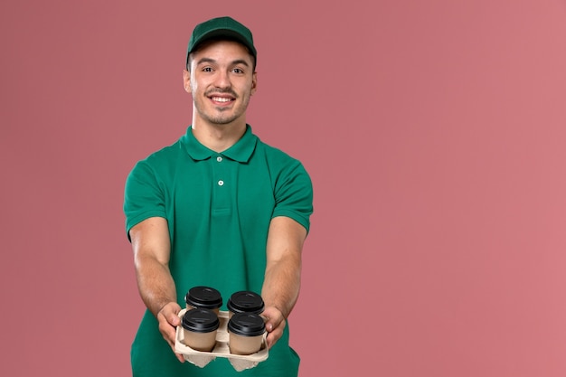 Mensajero masculino vista frontal en uniforme verde sosteniendo tazas de café de entrega entregándolas sobre fondo rosa