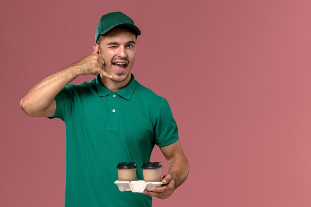 Mensajero masculino de vista frontal en uniforme verde sosteniendo tazas de café de entrega marrón y guiñando un ojo sobre fondo rosa