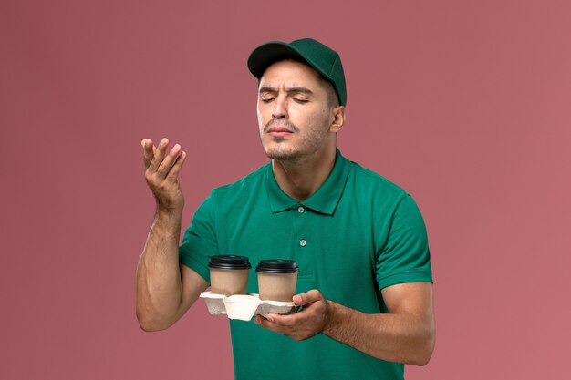 Mensajero masculino de vista frontal en uniforme verde sosteniendo tazas de café de entrega marrón oliendo su aroma sobre fondo rosa