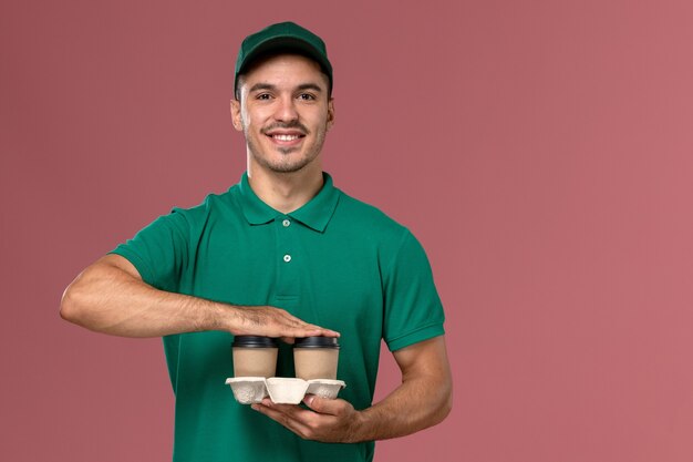 Mensajero masculino de vista frontal en uniforme verde sosteniendo tazas de café de entrega marrón y sonriendo sobre el fondo rosa