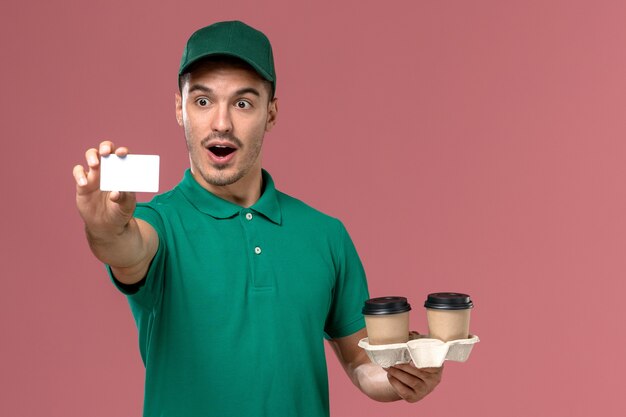 Mensajero masculino de vista frontal en uniforme verde sosteniendo tazas de café de entrega marrón y tarjeta blanca en el escritorio rosa macho