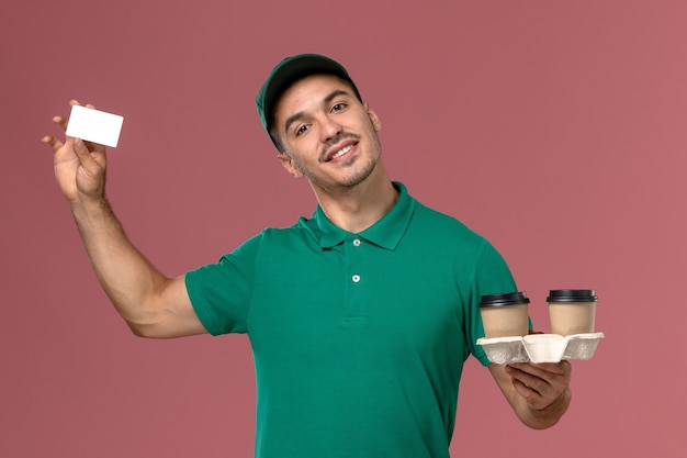 Mensajero masculino de vista frontal en uniforme verde sosteniendo tazas de café de entrega marrón y tarjeta blanca en el escritorio rosa