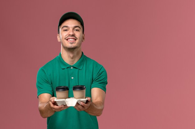 Mensajero masculino vista frontal en uniforme verde sosteniendo tazas de café de entrega sonriendo sobre el fondo rosa claro