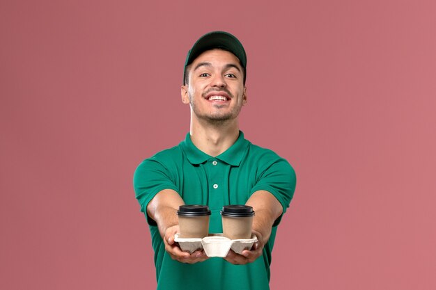 Mensajero masculino de vista frontal en uniforme verde sosteniendo tazas de café de entrega y sonriendo sobre fondo rosa