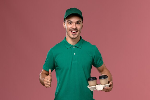 Mensajero masculino de vista frontal en uniforme verde sosteniendo tazas de café de entrega con sonrisa sobre fondo rosa