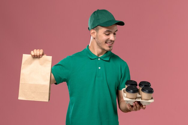 Mensajero masculino de vista frontal en uniforme verde sosteniendo tazas de café marrón y paquete de alimentos en el fondo rosa