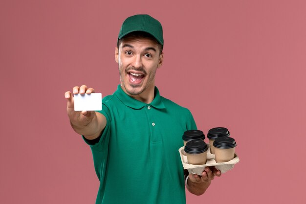 Mensajero masculino de vista frontal en uniforme verde sosteniendo tazas de café marrón y tarjeta de regocijo sobre fondo rosa