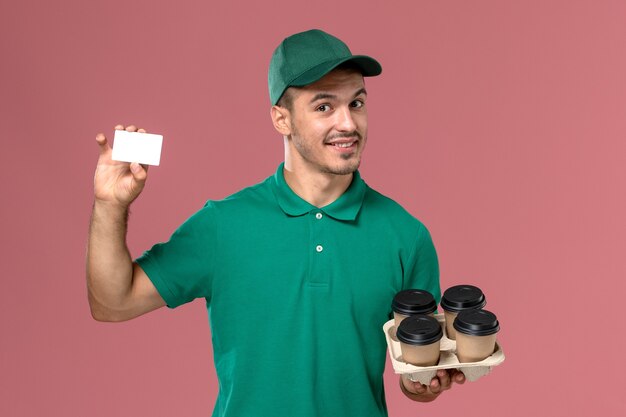 Mensajero masculino de vista frontal en uniforme verde sosteniendo tazas de café marrón y tarjeta sobre fondo rosa