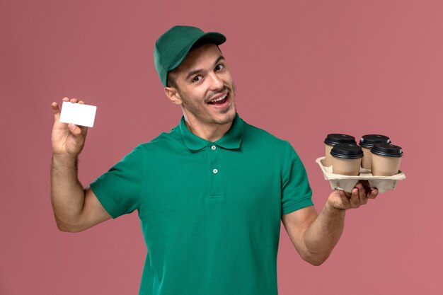 Mensajero masculino de vista frontal en uniforme verde sosteniendo tazas de café marrón y tarjeta sobre fondo rosa