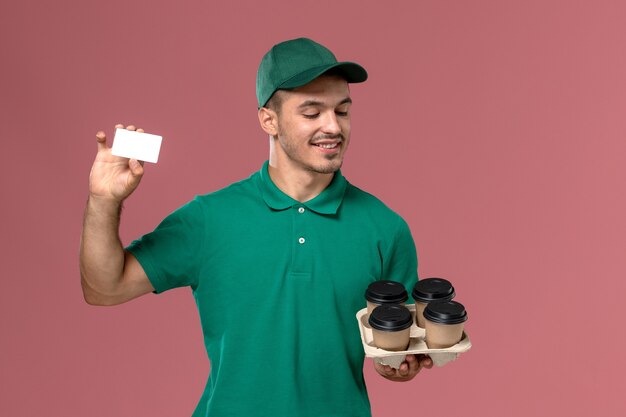 Mensajero masculino de vista frontal en uniforme verde sosteniendo tazas de café marrón y tarjeta sobre fondo rosa