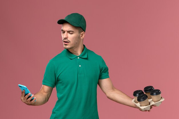 Mensajero masculino de vista frontal en uniforme verde sosteniendo tazas de café marrón con teléfono en el fondo rosa