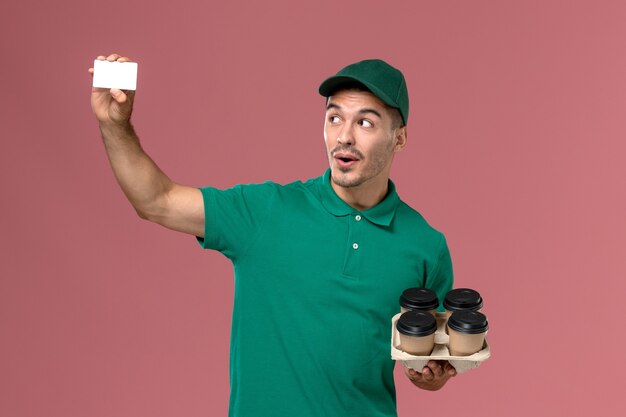 Mensajero masculino de vista frontal en uniforme verde con tazas de café marrón y tarjeta en el rosa