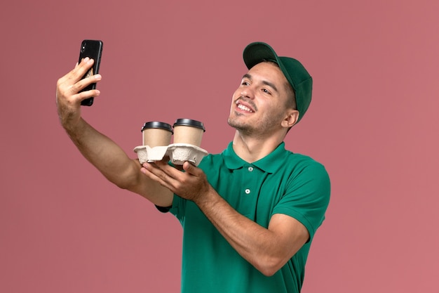Mensajero masculino de vista frontal en uniforme verde tomando una foto con café en el fondo rosa