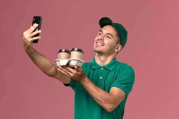 Mensajero masculino de vista frontal en uniforme verde tomando una foto con café en el fondo rosa