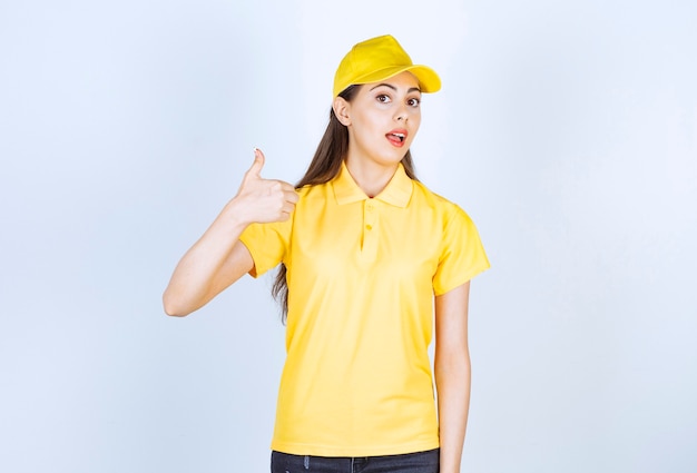 Mensajero de mujer hermosa en uniforme amarillo de pie y dando pulgar hacia arriba sobre la pared blanca.