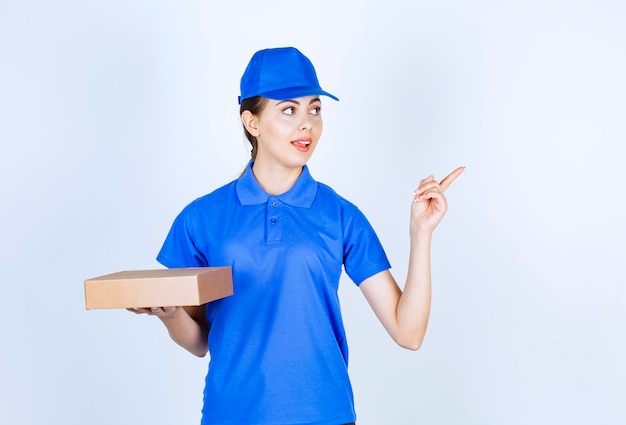 Mensajero mujer joven en uniforme azul posando con caja de cartón sobre fondo blanco.