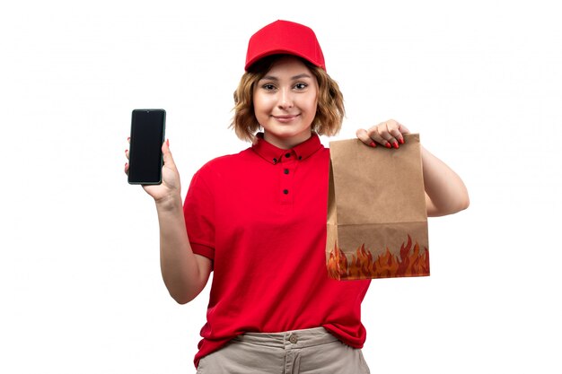 Un mensajero de mujer joven de vista frontal en camisa roja gorra roja sosteniendo el paquete de entrega y sonriendo smartphone