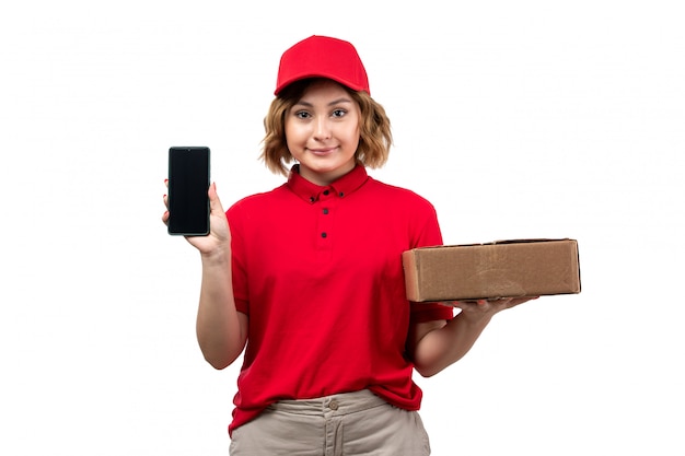 Foto gratuita un mensajero de mujer joven de vista frontal en camisa roja gorra roja sosteniendo el paquete de entrega y sonriendo smartphone
