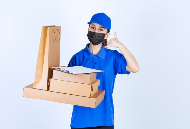 Mensajero mujer con máscara y uniforme azul sosteniendo una bolsa de cartón y varias cajas mientras pide una llamada.