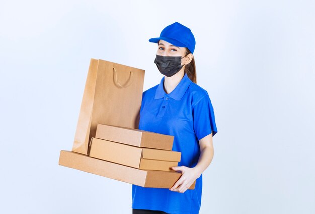 Mensajero mujer con máscara y uniforme azul sosteniendo una bolsa de cartón y varias cajas.