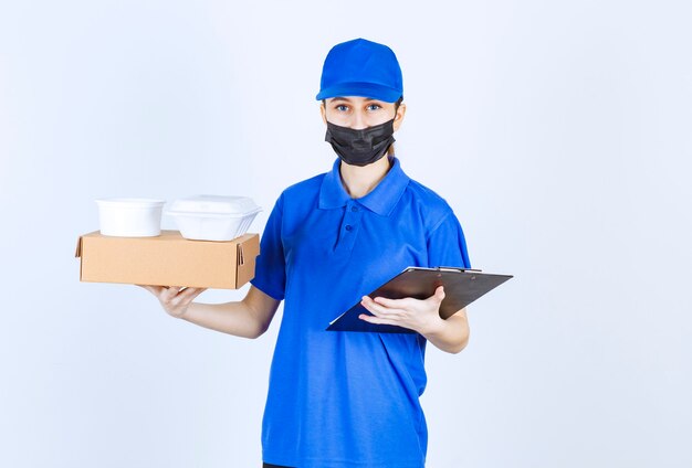 Mensajero mujer con máscara y uniforme azul sosteniendo una caja de cartón, paquetes para llevar y una carpeta negra.