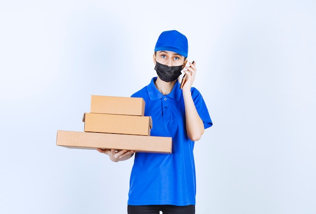 Mensajero mujer con máscara y uniforme azul sosteniendo un stock de cajas de cartón y hablando por teléfono.