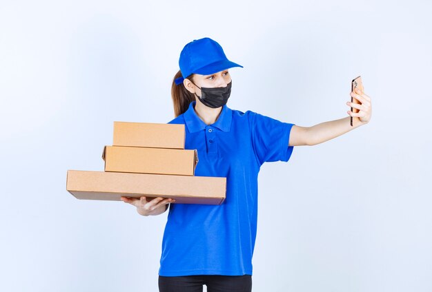 Mensajero mujer con máscara y uniforme azul sosteniendo un stock de cajas de cartón y haciendo una videollamada o tomando su selfie