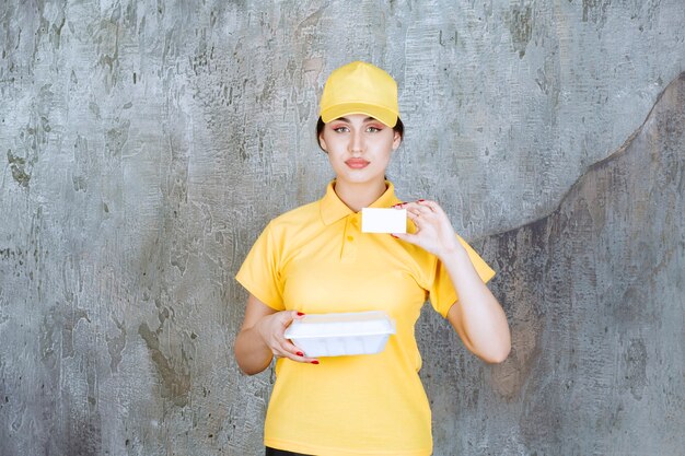 Mensajero mujer en uniforme amarillo entregando una caja de comida para llevar blanca y presentando su tarjeta de visita.