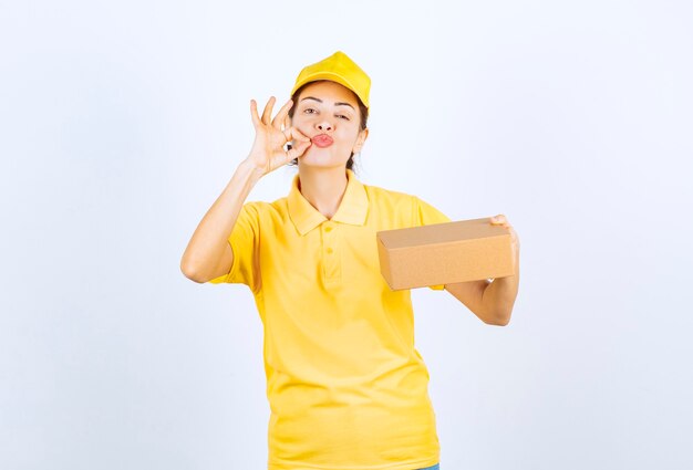 Mensajero mujer en uniforme amarillo entregando un paquete de cartón y mostrando un signo de mano exitoso.