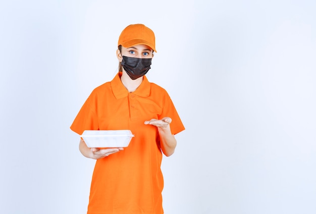 Mensajero mujer en uniforme amarillo y máscara negra sosteniendo una caja de comida para llevar de plástico y apuntando a ella.