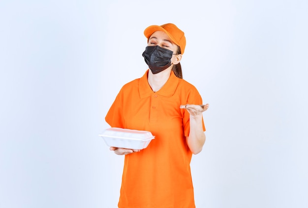 Mensajero mujer en uniforme amarillo y máscara negra sosteniendo una caja de comida para llevar de plástico y oliendo el sabor.