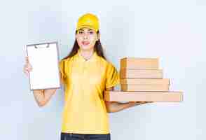 Foto gratuita mensajero mujer en uniforme amarillo con montón de cajas de cartón y portapapeles sobre pared blanca.