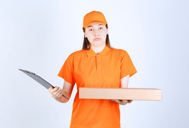 Mensajero mujer en uniforme amarillo sosteniendo una caja de cartón para llevar y comprobando la lista de direcciones y parece confundida.