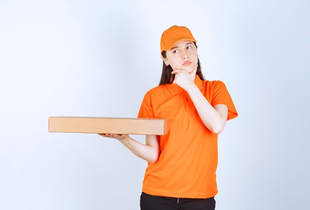 Mensajero mujer en uniforme amarillo sosteniendo una caja de cartón para llevar, parece confundido y pensando.