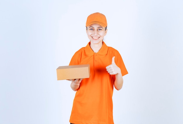 Mensajero mujer en uniforme amarillo sosteniendo una caja de cartón y mostrando un signo de mano positivo.