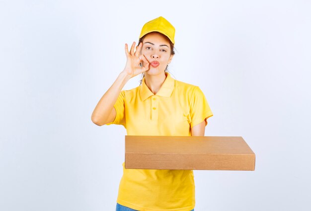 Mensajero mujer en uniforme amarillo sosteniendo un paquete de cartón y mostrando signo de mano de disfrute.