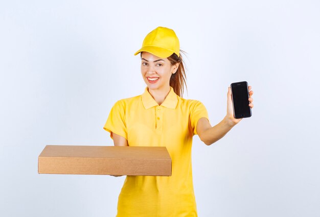 Mensajero mujer en uniforme amarillo sosteniendo un paquete de cartón y mostrando su teléfono inteligente negro.