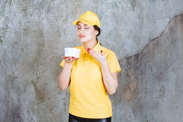 Mensajero mujer en uniforme amarillo sosteniendo una taza de comida para llevar y oliendo el producto.