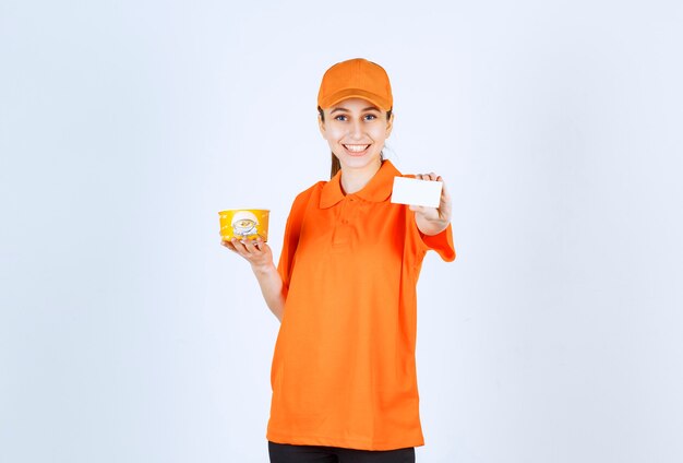 Mensajero mujer en uniforme amarillo sosteniendo una taza de fideos para llevar y presentando su tarjeta de visita.