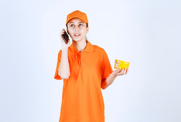 Mensajero mujer en uniforme amarillo sosteniendo una taza de fideos para llevar y tomando el pedido desde el teléfono inteligente.