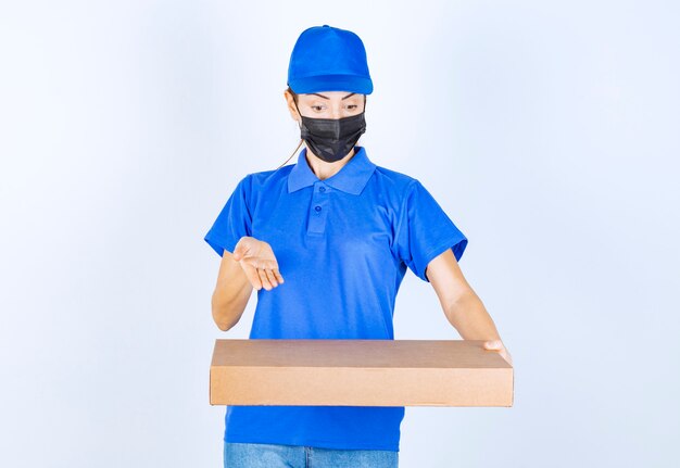 Mensajero mujer en uniforme azul y mascarilla entregando una caja de cartón al cliente.