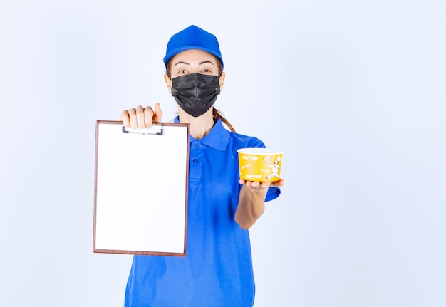 Mensajero mujer en uniforme azul y mascarilla entregando comida para llevar y pidiendo al cliente que firme.