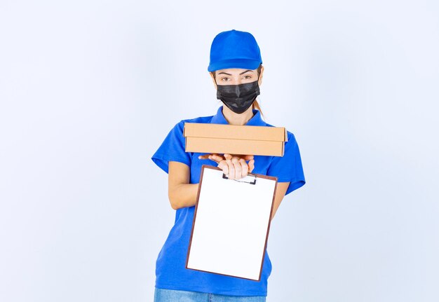 Mensajero mujer en uniforme azul y mascarilla entregando un paquete de cartón y pidiendo al cliente que firme en el espacio en blanco.