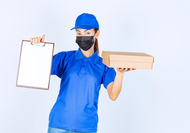 Mensajero mujer en uniforme azul y mascarilla entregando un paquete de cartón y pidiendo al cliente que firme en el espacio en blanco.