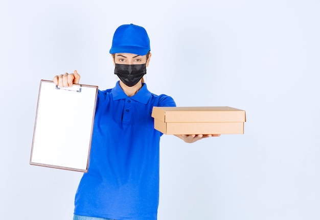 Mensajero mujer en uniforme azul y mascarilla entregando un paquete de cartón y pidiendo al cliente que firme en el espacio en blanco.
